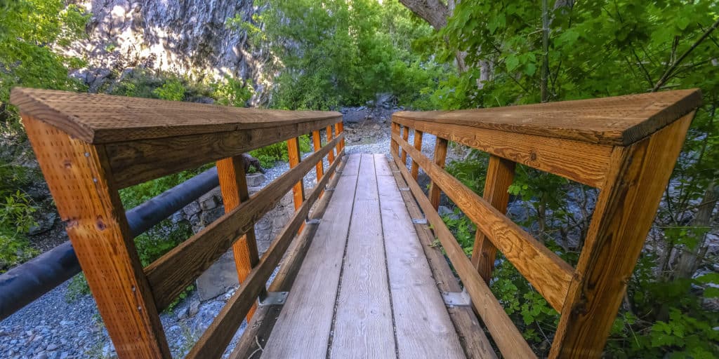 Bridge with rails on a rocky terrain in Provo. Narrow wooden bridge with rails on a hiking trail in Provo Canyon, Utah. The bridge leads to a rocky and rugged terrain with trees and lush foliage.
